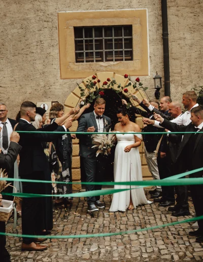 Braut und Bräutigam laufen durch einen Spalierbogen aus Rosen und Bändern – festlicher Hochzeitsmoment mit Gästen